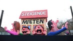 Ein Haus mit großer Aufschrift "Steigende Mieten", darunter sind mehrere Menschen eingequetscht