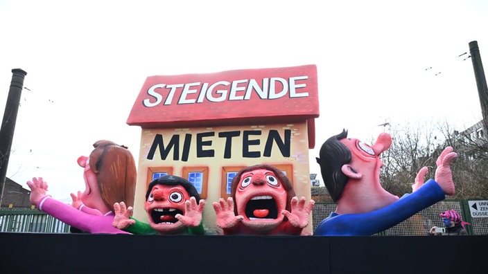 Ein Haus mit großer Aufschrift "Steigende Mieten", darunter sind mehrere Menschen eingequetscht