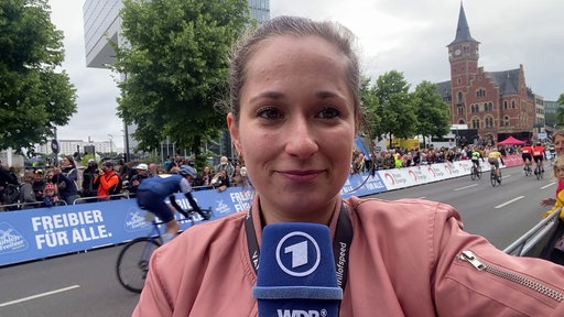Reporterin schaut in die Kamera, im Hintergrund fahren Radfahrer