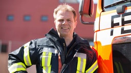 Der Pressesprecher Ulrich Laschet der Feuerwehr Köln lehnt an einem Feuerwehrwagen und lächelt in die Kamera