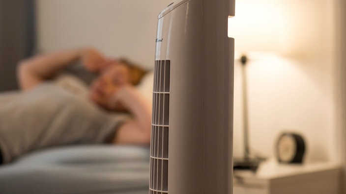 Eine Frau liegt im Bett, während ein Ventilator davor steht und die Luft kühlt