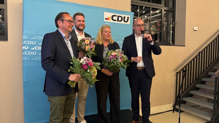 Vier Personen vor einem CDU- Banner. Drei halten einen Blumenstrauß. Die Person rechts spricht in ein Mikrofon. Ganz Links Obermürgermeister Thomas Kufen.