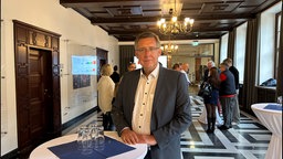 CDU Oberbürgermeisterkandidat Andreas Bracke am Stehtisch. Im Hintergrund eine Gruppe von Menschen und ein Bildschirm mit Wahlergebnissen. 