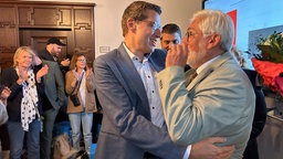 Bochums bisheriger OB Eiskirch umarmt den neuen OB Lukat. Im Hintergrund eine Gruppe Menschen und ein Bildschirm mit Wahlergebnissen. 