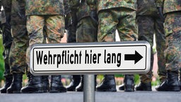 Ein Schild mit der Aufschrift "Wehrpflicht hier lang" steht vor Soldaten in Tarnuniform