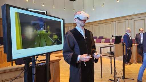 Gerichtsmitarbeiter im gerichtlichen Gewand trägt lächelnd VR-Brille, im Hintergrund läuft eine Straßenverkehrs-Simulation