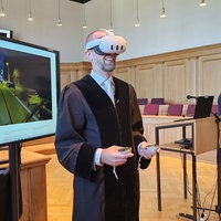 Gerichtsmitarbeiter im gerichtlichen Gewand trägt lächelnd VR-Brille, im Hintergrund läuft eine Straßenverkehrs-Simulation
