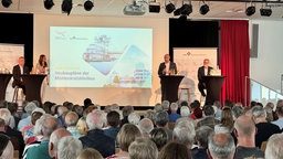 Vier Menschen bei einem Vortrag auf der Bühne. Ein Bild ist auf eine Leinwand gespielt.