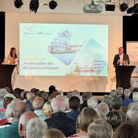 Vier Menschen bei einem Vortrag auf der Bühne. Ein Bild ist auf eine Leinwand gespielt.