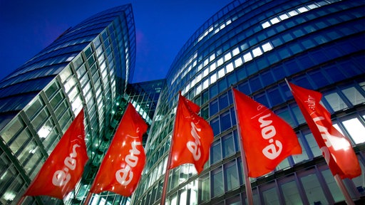 Die Zentrale von Eon in Essen: rote Fahnen mit Firmenlogo wehen vor einem hohen Gebäude mit beleuchteten Fenster vor dunkelblaumen Himmel
