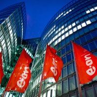 Die Zentrale von Eon in Essen: rote Fahnen mit Firmenlogo wehen vor einem hohen Gebäude mit beleuchteten Fenster vor dunkelblaumen Himmel