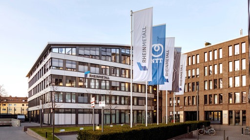 Mehrere Fahnen mit der Aufschrift "Rheinmetall" und Logo stehen vor einem modernen Bürogebäude mit Glasfassade.