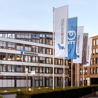 Mehrere Fahnen mit der Aufschrift "Rheinmetall" und Logo stehen vor einem modernen Bürogebäude mit Glasfassade.