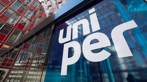 Großes blaues Schild mit weißer Aufschrift „uni per“ vor einer modernen Glas- und Stahlkonstruktion eines Gebäudes.