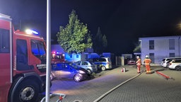 Zwei Feuerwehrmenschen stehen nachts vor einem Haus. Links ein Feuerwehrwagen, ein Wasserschlauch liegt auf der Straße