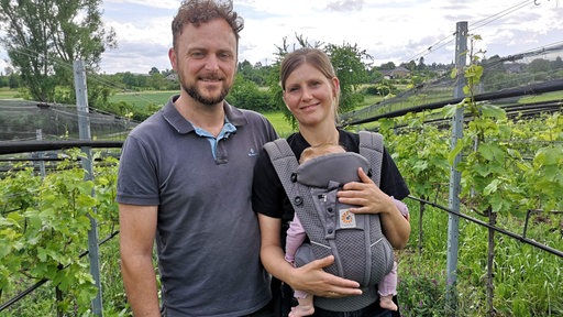 Ein Paar steht zusammen in seinem Weinberg und sie haben ein Baby dabei.