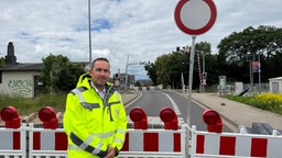 Ein Mann steht in gelber Warnjacke vor einer ABsperrung, neben ihm ein "DUrchfahrt Verboten"-Schild.