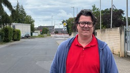 EIn Mann steht in roten Polo-Hemd und blauer Jacke an einer Straße. Er trägt einen Mittelscheitel und eine Politenbrille mit schwarzem Rand. 