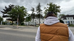 Ein Mann steht mit dem Rücken zur Wand vor einem Gebäude mit einem Zaun. Er trägt eine braune Weste und einen grauen Pullover.