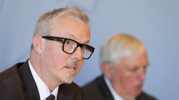 Ein Mann mit grauem Haar und Hornbrille erklärt auf einer Pressekonfernenz einen Sachverhalt.