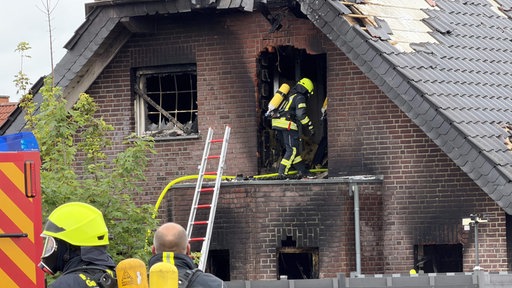 Ein Feuerwehrmann tritt in ein Backsteinhaus, das ein vom Brand zerstörtes Dach hat. Es hat schwarze Rußschleier an den Wänden.