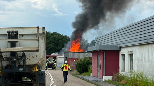 Auf dem Bild ist ein größerer Brand mit hohen Flammen und dichtem schwarzem Rauch zu sehen. Mehrere Personen in Warnkleidung befinden sich in der Nähe des Einsatzortes, während das Feuer im Hintergrund brennt.