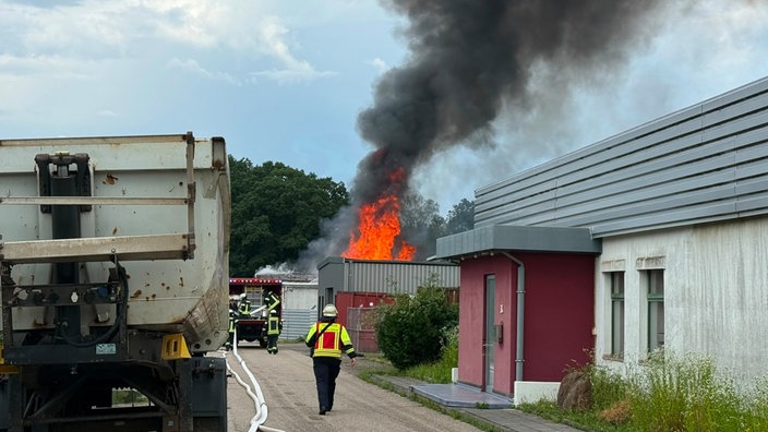 Auf dem Bild ist ein größerer Brand mit hohen Flammen und dichtem schwarzem Rauch zu sehen. Mehrere Personen in Warnkleidung befinden sich in der Nähe des Einsatzortes, während das Feuer im Hintergrund brennt.
