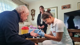 Eine junge Frau mit blauen Gummihandschuhen und weißer Kleidung sticht einem älteren Herren mit eine Lanzette in den Finger, um seinen Blutzucker zu messen. 