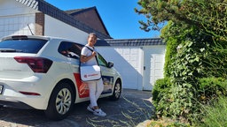Eine junge Frau steht lächelnd in einer Garagenauffahrt an einen Pkw gelehnt. 