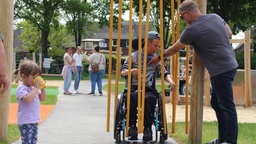 Auf einem Spielplatz spielt ein Junge im Rollstuhl mit einem Vorhang aus Stäben. Sein Vater steht daneben und hilft ihm, während ein kleines Mädchen im Vorgerdrung aus einem Becher trinkt. 
