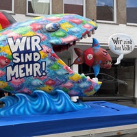 Ein großer Fisch mit der AUfschrift "wir sind mehr" isst einen kleineren Fisch auf dem "wir sind das Volk" steht.