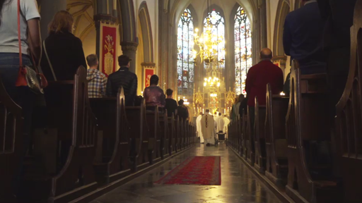 In einer Kirche steht eine Reihe Menschen in der Schlange um die Kommunion zu empfangen. Sie stehen vor dem Priester in liturgischen Gewändern und dem goldenen Altar 