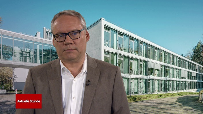 Ein Mann mit schwarzer Brille und grauem Jacket vor einem dreistöckigen Gebäude mit großen Glasfenstern. Unten Links das rote Logo der Aktuellen Stunde.