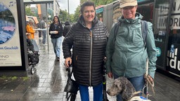 Eine Frau mit einem Rollator und eine Frau mit einem Assistenzhund für Blinde stehen vor einem Bus