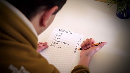 Ein Schüler sitzt über einem handgeschriebenen Wahlzettel und hat einen Stift in der Hand