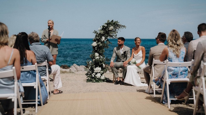 Ein Hochzeitspaar sitzt am Ufer eines türkisblauen Meeres. Neben ihnen steht eine Frau mit einer Mappe an einem Mirko. Hochzeitsgäste sitzen auf Stühlen und schauen aufs Paar. 