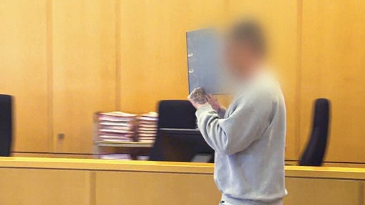 Ein Mann mit grauem Pullover in einem Gerichtssaal. Sein Gesicht ist verpixelt, er verdeckt es zusätzlich mit einem Aktenordner. 
