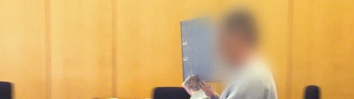 Ein Mann mit grauem Pullover in einem Gerichtssaal. Sein Gesicht ist verpixelt, er verdeckt es zusätzlich mit einem Aktenordner. 