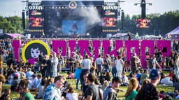Ein pinker Schriftzug zeigt das Wort "Pinkpop", drumherum zahlreiche Besucher auf der Wiese, dahinter die Bühne