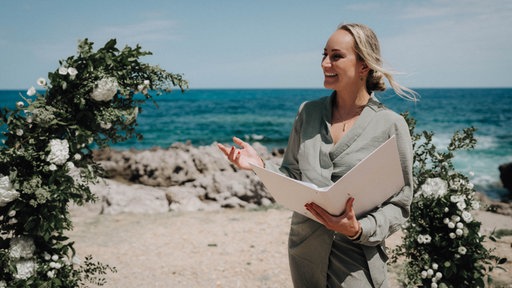 Samantha Meyer redet vor Strand-Kulisse zum Publikum einer Hochzeit