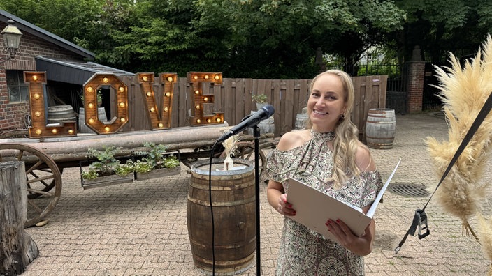 Eine blonde Frau im festlichen Sommerkleid steht mit einer aufgeschlagenen Mappe vor einem Mirko. Im Hintergrund ein Innenhof, der mit alten Holzfässern und einem "Love-"Schriftzug ebenfalls aus Holz dekoriert ist.