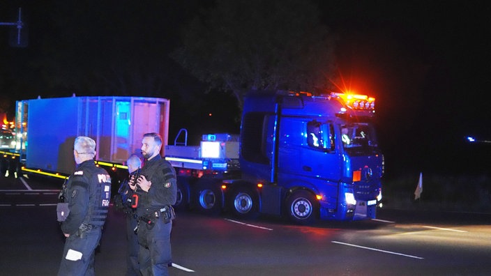 In der Dunkelheit fährt ein LKW mit einem Anhänger. Auf dem Anhänger ist ein weißer Castor-Behälter. Im Vordergrund stehen Polizisten.
