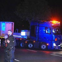 In der Dunkelheit fährt ein LKW mit einem Anhänger. Auf dem Anhänger ist ein weißer Castor-Behälter. Im Vordergrund stehen Polizisten.