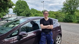 Eine Frau in Arbeitshose steht vor ihrem Auto