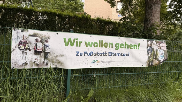 An einem Zaun hängt ein Banner mit der Aufschrift "Wir wollen gehen! Zu Fuß statt Elterntaxi. Kreis Mettmann".