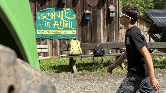 Eine Holzhütte, auf der Wand befindet sich eine Plane mit der Aufschrift "Schule in Arbeit". Auf der rechten Seite ein Jugendlicher mit Sonnenbrille.