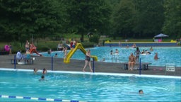 Menschen baden und entspannen in einem Freibad mit mehreren Becken und Wasserrutschen.