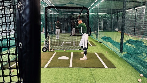 Man sieht einen Baseballspieler in grüner Spieluniform. Er hält einen Baseballschläger in der Hand und steht in einem Schlagkäfig.