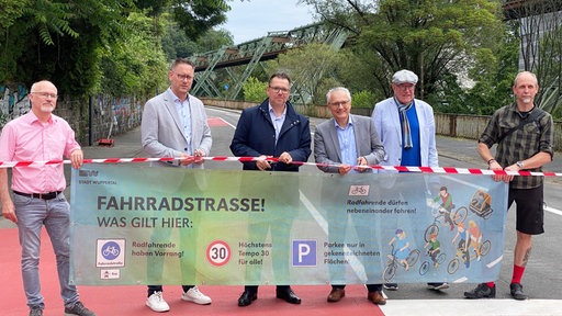 Sechs männliche Vertreter der Stadt Wuppertal halten einen Banner mit der Aufschrift Fahrradstrasse und Verkehrssymbolen, im Hintergrund das Schwebebahngerüst