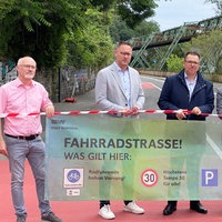 Sechs männliche Vertreter der Stadt Wuppertal halten einen Banner mit der Aufschrift Fahrradstrasse und Verkehrssymbolen, im Hintergrund das Schwebebahngerüst